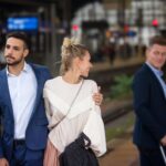 Stel op station, man slaat arm om vrouw, zij glimlacht naar andere man.